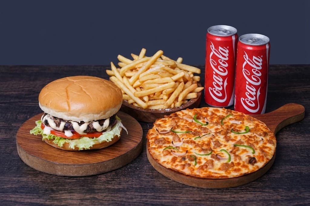 A burger, a bowl of fries and a pizza on wooden boards with two Coke cans